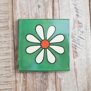 May include: A green ceramic tile featuring a white daisy flower with an orange centre. The tile is ideal for adding a touch of vintage charm to any space.