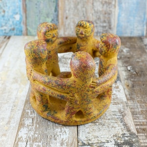 May include: A ceramic candle holder depicting five figures holding hands in a circle. The figures are painted yellow with red and green details. This decorative item is a unique piece of home decor, suitable for a table or shelf.