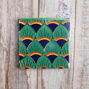 May include: Square ceramic tile featuring a repeating pattern of green, blue, and orange fan-shaped designs. The tile has a glossy finish and is set against a light-coloured wooden surface.