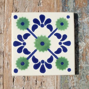 May include: A square ceramic tile with an off-white base colour featuring a symmetrical pattern. The design includes a central green star-shaped flower, four surrounding blue petal motifs, and four green star-shaped flowers at the corners.