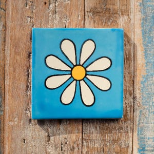 May include: Square ceramic tile featuring a daisy design on a bright blue background. The daisy has white petals with black outlines and a yellow centre. The tile is set against a rustic wooden surface.