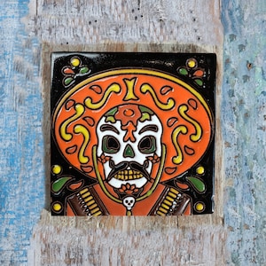 May include: A colourful ceramic tile depicting a Day of the Dead sugar skull wearing a sombrero. The skull is decorated with orange, yellow and black details. The sombrero is orange with black and yellow designs.