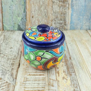 May include: A hand-painted ceramic jar with a lid. The jar is round with a dark blue rim and lid. The jar and lid are decorated with colourful floral and abstract patterns in shades of green, orange, blue, and red. The jar sits on a light-coloured wooden surface.