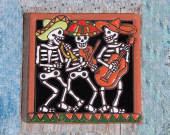 May include: Square ceramic tile featuring a Day of the Dead band. Three skeleton musicians in colorful sombreros play a trumpet, violin, and guitar against a black background. The tile has a brown border and a green and orange patterned base.