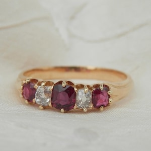 Op de afbeelding: Een gouden ring met vijf edelstenen: twee rode robijnen, twee heldere diamanten en een centrale robijn. De ring heeft een gepolijste gouden band en de edelstenen zijn ovaal geslepen, in een rij geplaatst. De ring wordt getoond op een wit, getextureerd oppervlak.