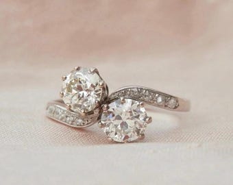 May include: A platinum ring featuring two large, round-cut diamonds. The ring's band curves gracefully, set with smaller accent diamonds. The diamonds sparkle against a soft, neutral background, showcasing the ring's elegant design.