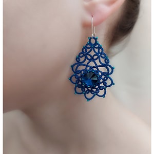 May include: A close-up of a blue, handmade earring. The earring has a detailed, lacy design with a large, dark blue crystal at the centre. The earring is suspended from a silver hook. The Instagram handle "@nonna_sergeeva" is visible.