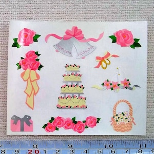 May include: Two sheets of wedding-themed stickers. One sheet features purple line drawings of wedding elements, including a cake, gift, and invitation. The other sheet has pink roses, a tiered cake, and bells. A ruler shows the sheets are about 3 inches wide.