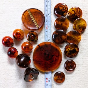 May include: A collection of vintage buttons in various sizes and shapes, arranged on a white surface. The buttons are primarily shades of brown and amber, with a ruler providing scale. The largest button is approximately 2 inches in diameter.