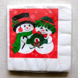 May include: A stack of white paper napkins with a festive Christmas design. The design features two snowmen with top hats, scarves, and a wreath, set against a red background with snowflakes.
