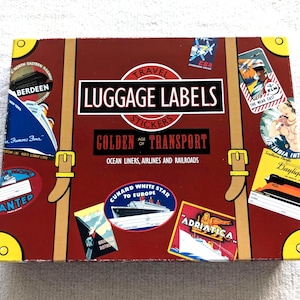 May include: A vintage-style box of travel luggage labels. The box is a deep red with faux leather straps and gold-colored buckles. The labels feature images of ocean liners, airplanes, and railroads, with text that reads "LUGGAGE LABELS" and "GOLDEN AGE OF TRANSPORT."