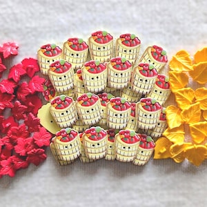 May include: A collection of small craft buttons featuring red maple leaf shapes, yellow maple leaf shapes, and cream-colored bushel baskets filled with red apples. The buttons are arranged in three distinct groups on a plain white surface.