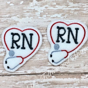 Può includere: Due toppe di feltro a forma di cuore rosso con sfondo bianco e uno stetoscopio grigio. Il testo "RN" è ricamato in nero su ogni toppa.