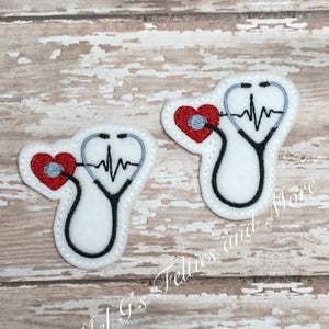 Può includere: Due toppe di feltro bianco con cuori rossi ricamati e stetoscopi neri e grigi. Gli stetoscopi hanno un cuore rosso all'estremità e una linea ECG nera e grigia. Il testo "Mel G's Felties and More" è visibile nell'angolo in basso a destra.