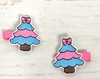 Puede incluir: Dos pinzas para el pelo de árbol de Navidad de color rosa y azul con un lazo rosa en la parte superior. Las pinzas están sobre un fondo de madera blanco.