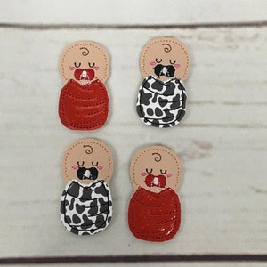 Puede incluir: Cuatro aplicaciones de bebé de fieltro, dos con estampado de vaca rojo y blanco y dos con tela de brillo rojo. Cada aplicación tiene un chupete rosa y los ojos cerrados.