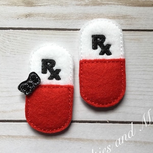 Puede incluir: Dos formas de píldoras de fieltro rojo con tapas blancas y letras negras "Rx". Una píldora tiene un lazo negro en el lateral.