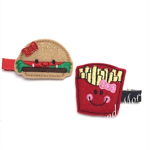 May include: Two felt food hair clips. One is a brown hamburger with a red tomato, lettuce, and a pink bow. The other is a red french fry container with a yellow french fry and a pink bow.