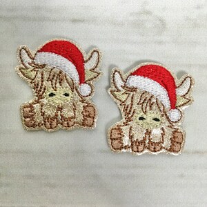 May include: Two embroidered patches of a brown Highland cow wearing a red Santa hat. The patches are on a white wooden background.