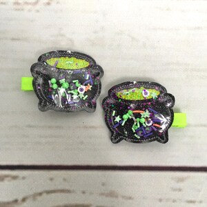 Pode incluir: Dois clipes de cabelo em forma de caldeirão preto com glitter verde e várias confeitas com tema de Halloween dentro. Os clipes estão em um fundo de madeira clara.