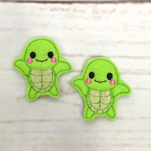 May include: Two green felt appliques of cartoon turtles with pink cheeks and black eyes. The turtles have a light green shell with a beige outline.