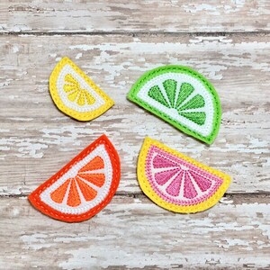 Op de afbeelding: Vier geborduurde vilt fruitplakjes, elk een ander citrusfruit: een gele citroen, een groene limoen, een oranje sinaasappel en een roze grapefruit. De plakjes zijn gerangschikt op een witgeschilderd houten oppervlak.