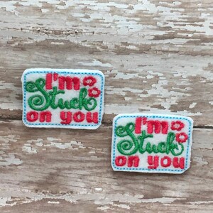 以下が含まれることがあります： 青いステッチが縁取りされた白いフェルト生地の2つのパッチ。各パッチには、「I'm stuck on you」という文字が緑と赤の刺繍で施されています。