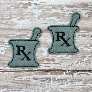 May include: Two felt appliques of a mortar and pestle with the letter "Rx" in the centre. The appliques are a light green colour with black stitching.