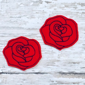 以下が含まれることがあります： 黒い輪郭のある赤いフェルトのバラが2つ。バラは白い木製の背景の上にあります。