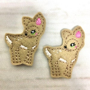 以下が含まれることがあります： ピンクの耳と黒い目をした、茶色のフェルト地の鹿のアップリケが2つ。鹿は白いディテールで刺繍されています。