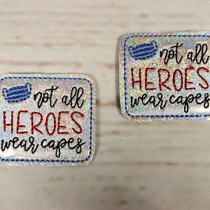 Puede incluir: Dos parches de planchado de purpurina plateada con costuras azules. Los parches son de forma cuadrada y presentan un gráfico de mascarilla facial azul con el texto "not all HEROES wear capes" en letras negras y rojas.