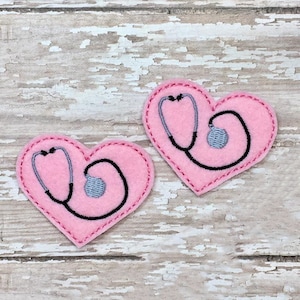 Puede incluir: Dos corazones de fieltro rosa con contornos bordados en negro. Cada corazón tiene un estetoscopio bordado en negro con un círculo gris para la pieza de escucha.