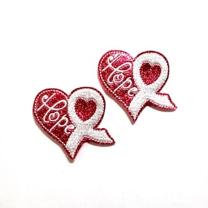 以下が含まれることがあります： 白いリボンと「Hope」という刺繍が施された、ピンクのグリッターハート型の2つのパッチ