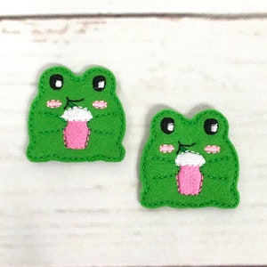 Könnte beinhalten: Zwei grüne Filz-Frosch-Applikationen mit schwarzen Augen, rosa Wangen und einer rosa-weißen Tasse in ihren Händen.