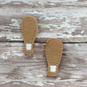 Puede incluir: Dos formas de zapatos de fieltro marrón con detalles de fieltro blanco. Los zapatos están cosidos alrededor de los bordes.