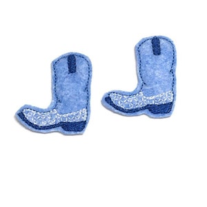 May include: Two light blue felt cowboy boot patches. Each boot has white embroidered details on the toe and a dark blue trim. The boots are set against a white background.