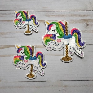 May include: Three embroidered iron-on patches featuring a white unicorn with a rainbow mane and tail on a carousel pole. The unicorn is facing left and has its eyes closed. The patches are different sizes.