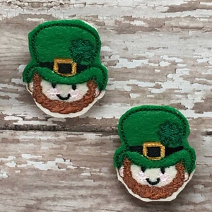 以下が含まれることがあります： 緑の帽子に金のバックルとクローバーが付いた、レプリコーンの頭のフェルトアップリケが2つ。レプリコーンは茶色のひげを生やし、笑顔をしています。