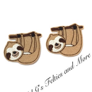 Puede incluir: Dos apliques de fieltro marrón en forma de perezoso con ojos negros y caras blancas, colgando de una rama de fieltro marrón. Los apliques miden aproximadamente 5 cm.