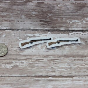 May include: Two small embroidered patches of rifles on a white background. The patches are about the size of a US quarter.