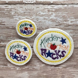 Pode incluir: Três círculos de feltro com costuras amarelas ao redor das bordas. O círculo maior diz "Kinder Garden Rocks" com uma maçã vermelha, estrelas roxas e costuras amarelas. Os dois círculos menores dizem "Kinder Garden Rocks" com uma maçã vermelha e estrelas roxas.