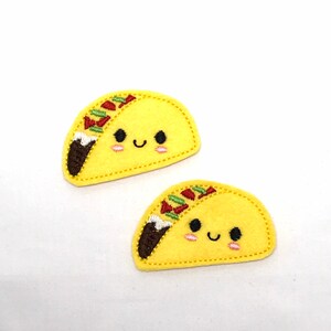 May include: Two yellow felt taco appliques with embroidered details, including a smiling face, and a brown, white, and green filling.