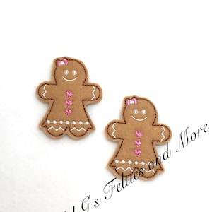 May include: Two brown felt gingerbread people with pink bows and pink hearts. The gingerbread people are embroidered with white details.
