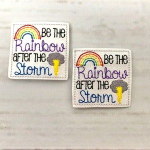 以下が含まれることがあります： カラフルな刺繍の虹と「Be the Rainbow after the Storm」というテキスト、そして黄色の稲妻のある灰色の雲が描かれた白い布のパッチが2つあります。