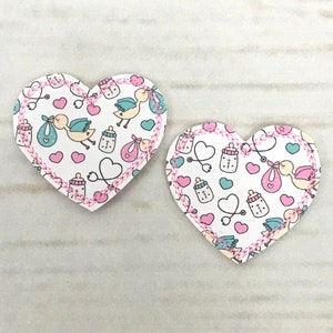 Puede incluir: Dos parches de tela en forma de corazón rosa y blanco con un patrón de temática infantil. El patrón incluye cigüeñas, biberones, corazones y estetoscopios.