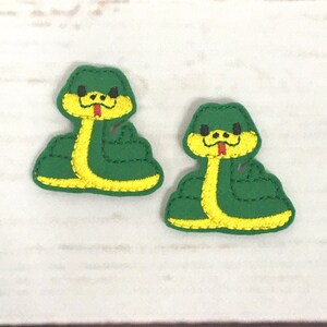 May include: Two green felt snake appliques with yellow stitching and red tongues. The snakes have black eyes and are coiled in a curled position.