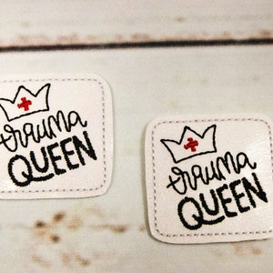 May include: Two white square patches with black stitching around the edges. Each patch features a red and white crown with a red cross in the center and the text "Trauma Queen" in black cursive lettering.