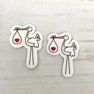Puede incluir: Dos parches bordados de cigüeña blanca con corazones rojos. Cada cigüeña lleva una pequeña bolsa blanca con un corazón rojo dentro.