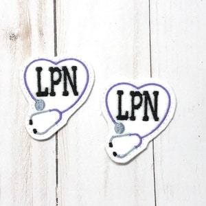 Può includere: Due toppe a forma di cuore in feltro bianco con cuciture viola. Ogni toppa ha uno stetoscopio ricamato in nero e il testo "LPN" in nero.