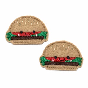 May include: Two felt appliques of a cartoon hamburger with a smiling face. The hamburger is brown with a red patty, green lettuce, and a white sesame seed bun.